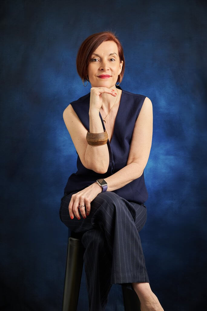 Femme assise sur tabouret haut, jambes et bras croisés, souriante portant une tenue bleue sur fond bleu
