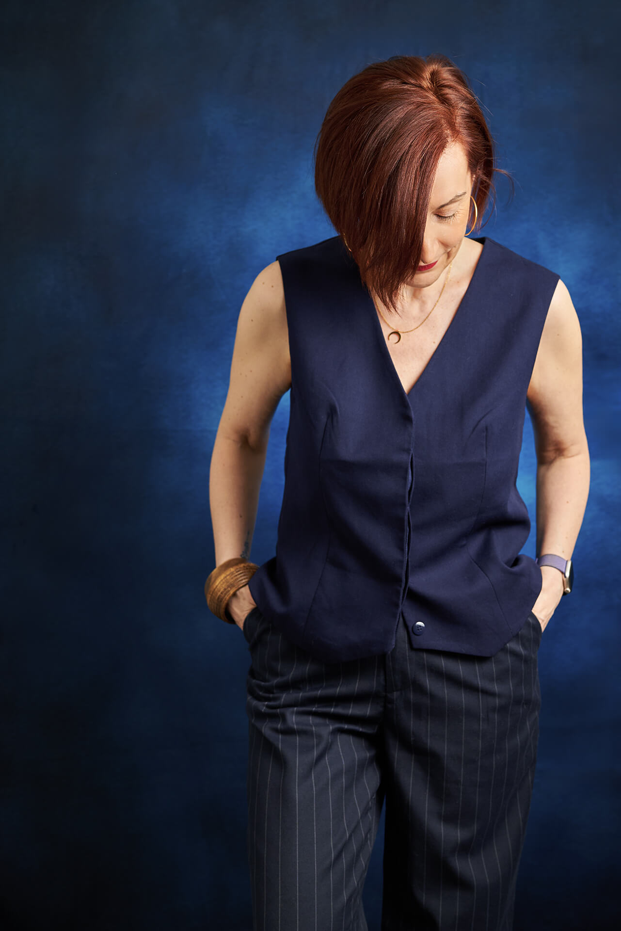 Femme tête baissée regardant sa tenue, elle porte un pantalon de costume rayé bleu et un gilet de costume bleu, elle est photographiée sur un fond bleu